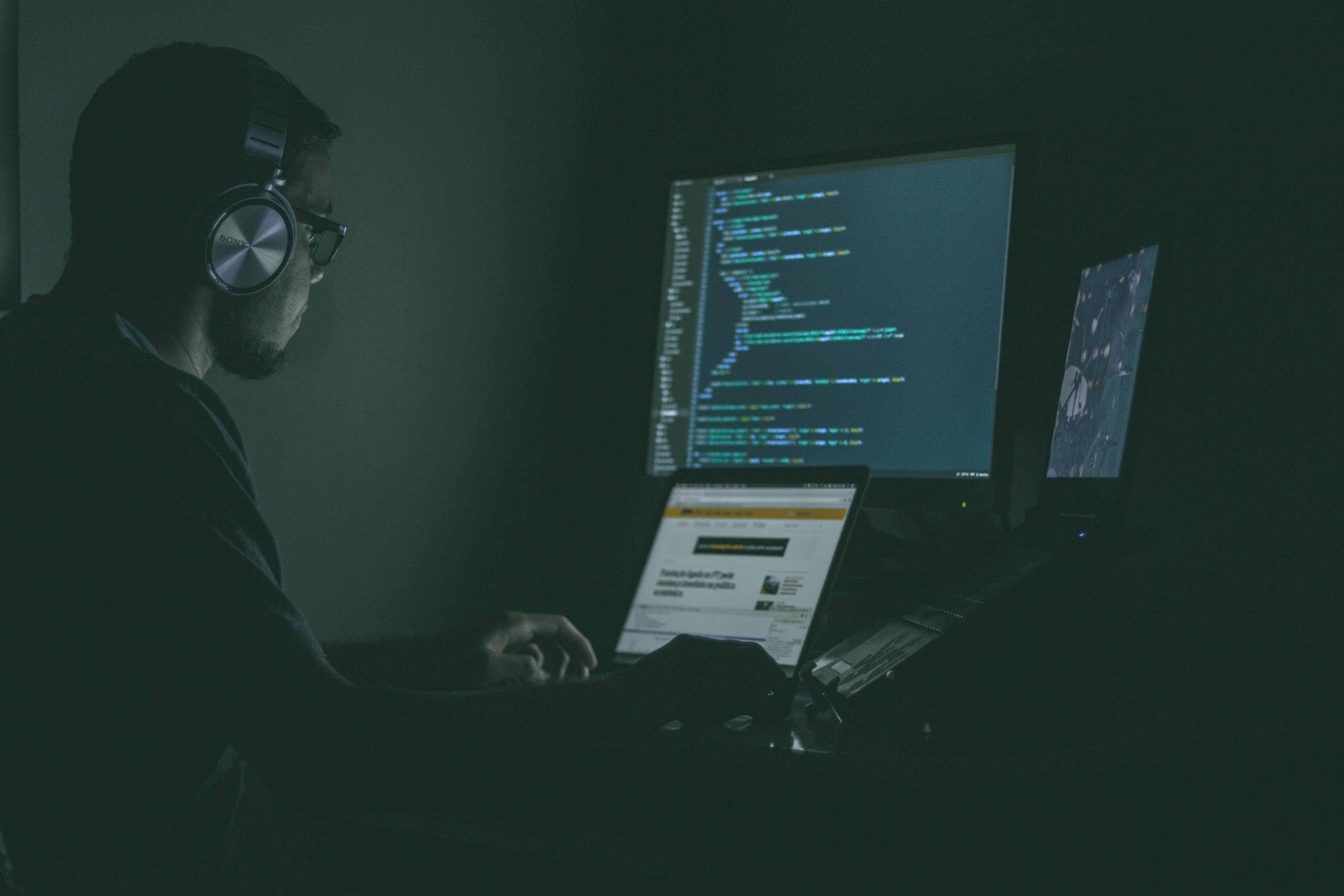A man wearing headphones sitting in the dark at his desk with three screens on it